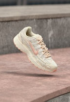 Beige and light pink Nike sneaker with mesh and synthetic upper, floating above brown concrete steps in outdoor setting.
